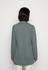 Capospalla grigio a maniche lunghe, con struttura testurizzata, colletto e taglio dritto, visto da dietro. Presenta una leggera curva sull'orlo.