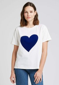 T-shirt blanc avec un grand motif de cœur bleu marine. Manches courtes, col rond, coupe droite. Porté avec un jean bleu. Tissu en coton lisse.