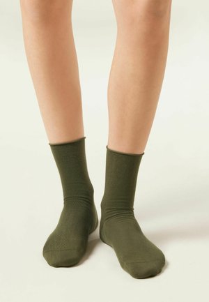 Jambes portant des chaussettes mi-mollet unies vert olive, debout sur une surface de couleur claire avec un fond neutre.