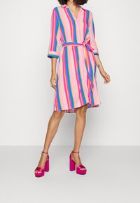 Robe rayée en rose, bleu et jaune avec un devant à boutons, un décolleté en V, et une ceinture à nouer à la taille. Elle possède des manches trois-quarts.