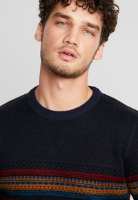 Navy gestrickter Pullover mit strukturiertem Körper und einem gemusterten Streifen in Rot, Orange und Blau über der Brust. Klassischer Rundhalsausschnitt.