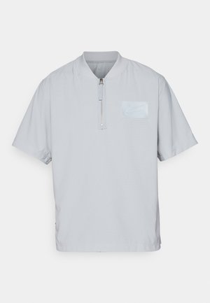 Polo-shirt à demi-zip gris clair avec manches courtes, présentant un tissu texturé et un patch logo subtil sur la poitrine.