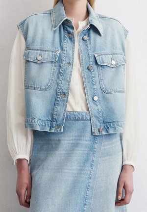 Nainen pukeutuneena vaaleansiniseen farkkutakkiin ja vastaavaan hameeseen, valkoinen pitkähihainen paita alla, seisoo valkoisen seinän edessä.