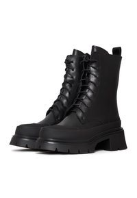 Botas de tobillo de cuero negro con cordones, suela de plataforma gruesa, puntera redondeada y detalles acanalados; tirador en la parte trasera para facilitara la colocación.