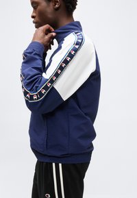 Hombre cerrando con cremallera una chaqueta deportiva azul marino y blanca con rayas con patrón de logos en las mangas, vistiendo pantalones negros con doble raya blanca lateral.