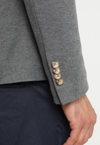 Blazer gris texturé avec une manche à quatre boutons dans des tons marron clair, doté d'un design épuré et d'une finition en tissu lisse.