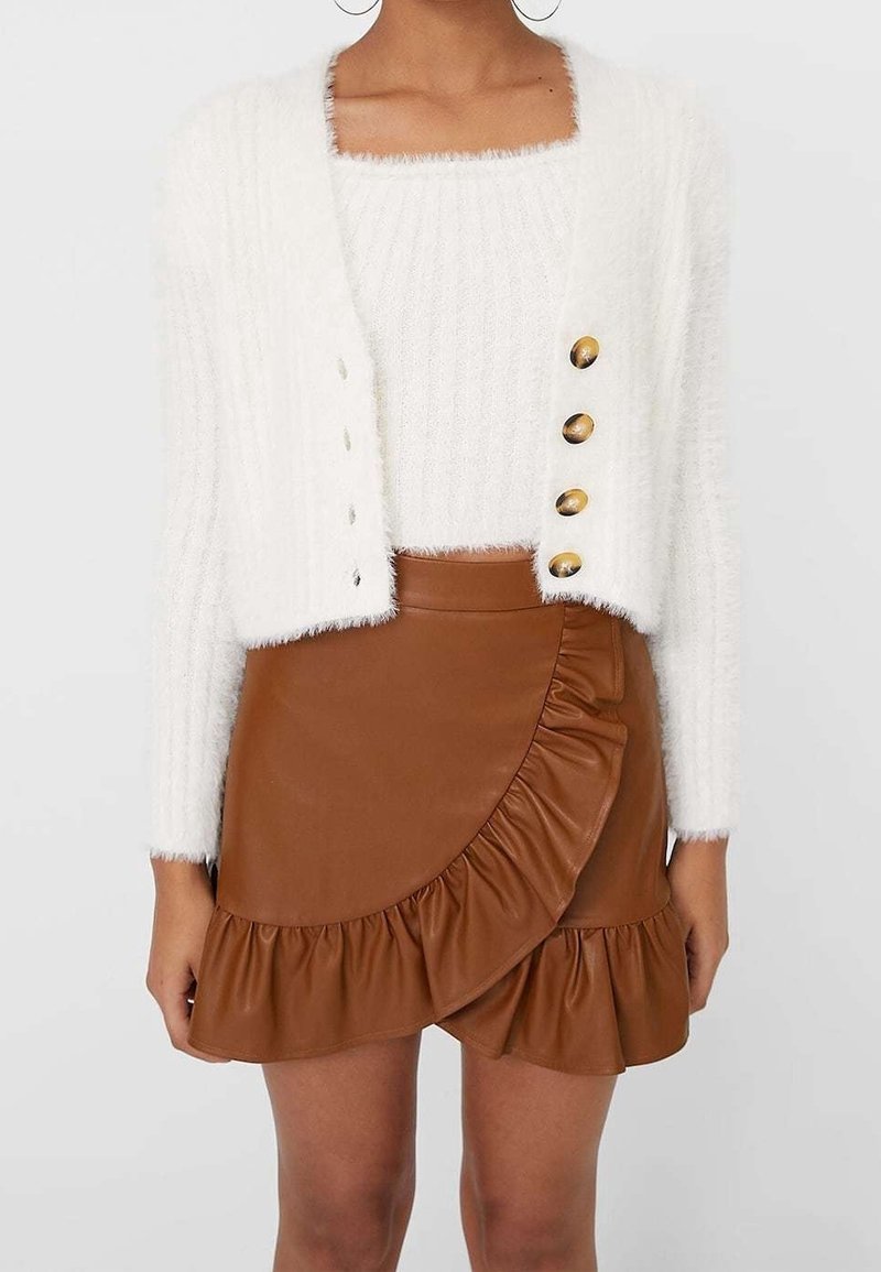 Cardigan court blanc duveteux avec boutons dorés sur un top blanc duveteux assorti, accompagné d’une mini-jupe marron en similicuir à volants.