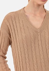 Beiger Strickpullover mit V-Ausschnitt, Zopfmuster und gerippten Akzenten am Kragen und an den Ärmeln.