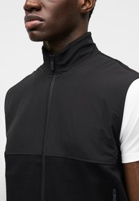 Μαύρο αμάνικο γιλέκο με ψηλό γιακά, φερμουάρ μπροστά και αντίθετη υφασμάτινη επιφάνεια στο κάτω μέρος. Λείο φινίρισμα υφάσματος.