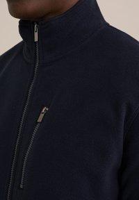 Tumesinine fleece-jakk kõrge kaelusega, millel on tõmblukuga rinnatasku ja tekstuurne kangas. Tõmbluku sulgemine metallist tõmbluku tõmblukuga.