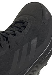 Gros plan sur une chaussure de sport noire avec un tissu texturé, trois bandes diagonales sur le côté, des lacets noirs et une semelle en caoutchouc nervurée.