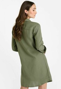 Robe chemise vert olive à manches longues, avec des poignets boutonnés et une texture lisse. Tombe au milieu de la cuisse avec un ourlet légèrement incurvé et un détail au dos.