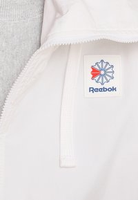 Weiße Zip-Jacke mit strukturiertem grauem Innenfutter, Reebok-Logo-Patch auf der Brust und strukturiertem Kordelzug-Design.