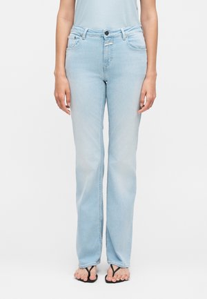 Γυναίκα που φοράει ανοιχτά μπλε τζιν με ίσια μπατζάκια και μαύρα σανδάλια τύπου φλιπ-φλοπ, στέκεται μπροστά σε ένα απλό λευκό φόντο.