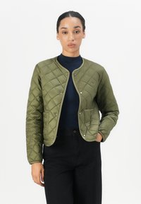 Olive green quilted jacket with a round collar, snap buttons, and two front pockets; paired with a dark turtleneck and black trousers.