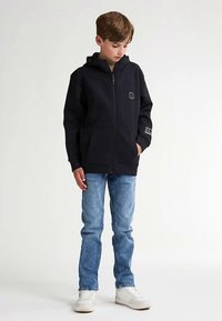 Schwarzer Reißverschluss-Hoodie mit ausgeprägter Kapuze, vorderen Taschen und rundem Logo auf der Brust. Kombiniert mit hellblauen Jeans und weißen Sneakers.