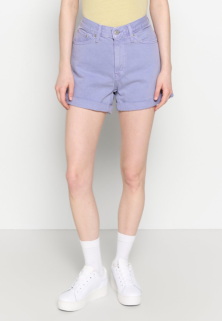 Paarse spijkerhotpants met opgerolde mouwen en voorkruiszakken, gecombineerd met een lichtgele top en witte sneakers met dikke zolen.