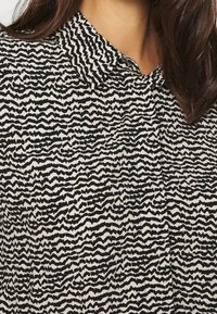 Blouse à motifs noir et blanc avec un design en zigzag ondulé, col classique et fermeture par boutons. Tissu texturé avec une finition douce.
