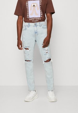 Lichtblauwe ripped jeans met rafelende hem en versleten details, gecombineerd met een donkerbruine graphic t-shirt en witte sneakers.