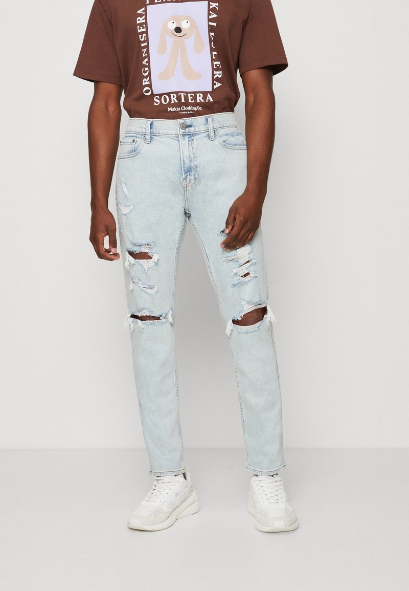 Ljusblå rivna jeans med fransade kanter och slitna detaljer, kombinerat med en mörkbrun t-shirt med tryck och vita sneakers.