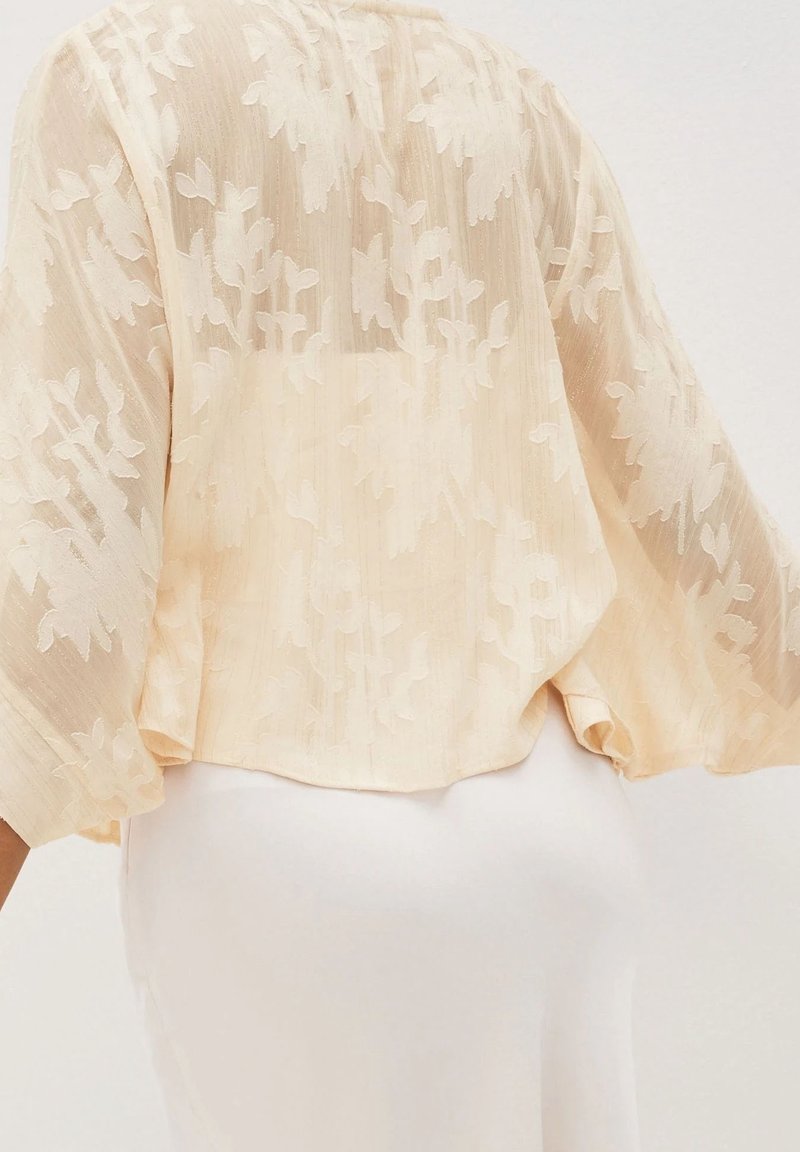 Blouse beige transparente à motif jacquard floral et manches larges portée sur une jupe ou une robe blanche vue de dos.