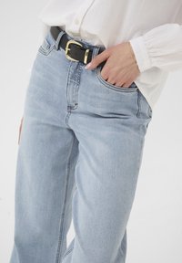 Frau, die eine hellblaue Jeans und ein weißes Shirt trägt, mit einer Hand in der Tasche, schwarzem Gürtel und goldener Armband am Handgelenk.