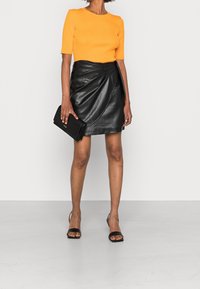 Oranje geribbelde top met korte mouwen, zwarte faux leren wikkelrok, zwarte clutch en zwarte sandaaltjes met bandjes op een lichtgrijze achtergrond.