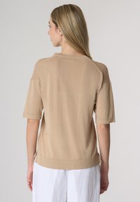Maglione beige lavorato a maglia con maniche corte, scollatura a coste e spacchi laterali. Texture liscia con vestibilità rilassata. Mostrato da dietro.