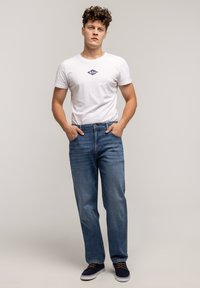 Biała bawełniana koszulka z granatowym logo, zestawiona z niebieskimi dżinsami. Model nosi również ciemne buty. Codzienny strój na neutralnym tle.