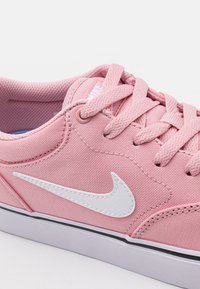 Rosa canvas Nike-sneaker med vit sula, vit swoosh-logga och rosa snören, som visar detaljer från sidan och ovanifrån.
