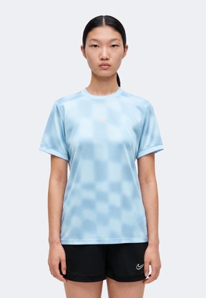 Une femme porte un haut de sport bleu clair à manches courtes et à motifs, ainsi qu’un short de sport noir, sur un fond uni.