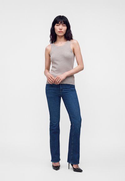 Jeune femme aux longs cheveux foncés portant un haut gris sans manches, un jean droit bleu et des chaussures à talons noires, debout devant un fond uni.