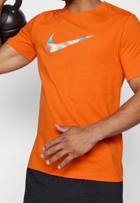 Camiseta de algodón naranja con un colorido logo de Nike en el pecho, diseño de cuello redondo y mangas cortas. Detalle de tejido texturizado visible.