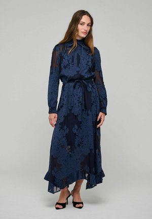 Donna che indossa un lungo abito di pizzo floreale blu navy con maniche lunghe trasparenti e sandali neri con tacco alto, in piedi su uno sfondo semplice.
