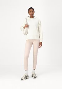 Sudadera capucha crema con un corte relajado, combinada con pantalones ajustados rosa claro y zapatillas blancas con detalles en negro. Lleva una bolsa beige.