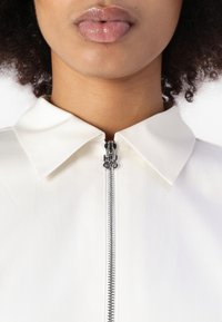 Camisa blanca con cremallera y cuello puntiagudo, con un zipper plateado que presenta un tirador cromado marcado con "HUGO". Textura de tela suave.