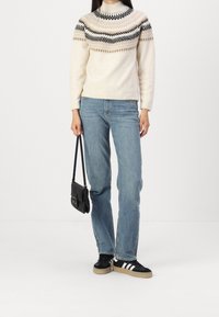 Strickpullover in Creme mit schwarzen und taupefarbenen geometrischen Mustern, kombiniert mit hellblauen Jeans und schwarzen Turnschuhen mit weißen Streifen.