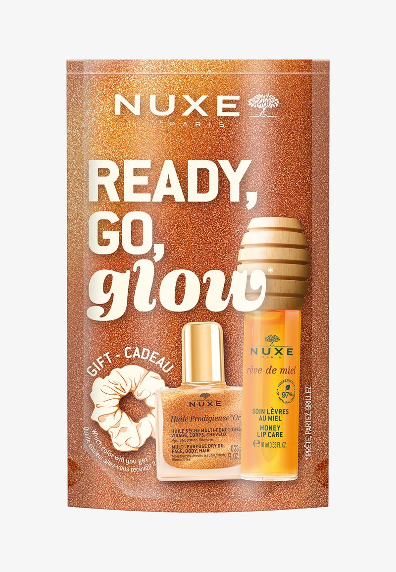 Zestaw prezentowy NUXE z błyszczącym, brązowym opakowaniem, butelką złotego oleju, tubką do pielęgnacji ust z miodem oraz gumką do włosów.
