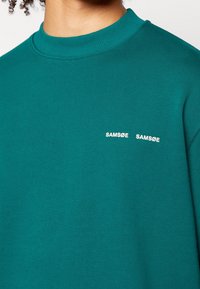 Modrý svetr s žebrovaným výstřihem a dvěma bílými logy "SAMØE" na hrudi. Měkká látka s hladkou texturou a pohodlným střihem.