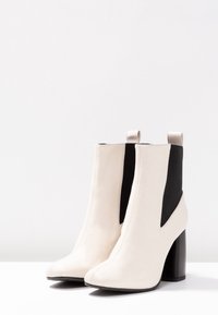 Bílé kožené kotníkové boty s černými elastickými panely, zaoblenou špičkou a čtveratým vysokým podpatkem. Hladký povrch s detaily šití.