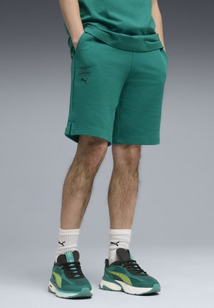 Jambes d'homme portant un short Puma bleu sarcelle avec le logo de l'équipe Aramco F1, des chaussettes blanches Puma et des baskets Puma bleu sarcelle, noir et jaune, debout sur un fond gris.