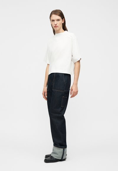 Γυναίκα που στέκεται φορώντας λευκή μπλούζα με κοντά μανίκια, σκουρόχρωμα τζιν με μανσέτες και πλαϊνές τσέπες, καθώς και μαύρα παπούτσια, μπροστά από έναν απλό φωτεινό φόντο.