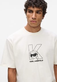 Krémové bavlnené tričko s čiernou grafikou postavy so slnečnými okuliarmi a veľkým písmenom "K" vedľa mena "KARL LAGERFELD."