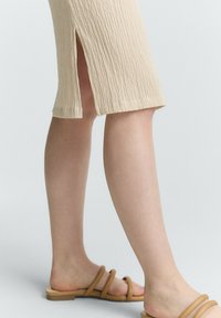 Abito beige a costine con spacco laterale, lunghezza al ginocchio. Indossato con sandali marroni con cinturini. Tessuto liscio e design minimalista.