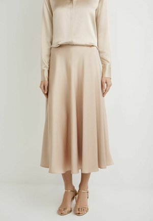 Kobieta w beżowej satynowej bluzce z długim rękawem i dopasowanej spódnicy midi, ubrana w złote sandały na wysokim obcasie z paskami, stojąca na jednolitym tle.