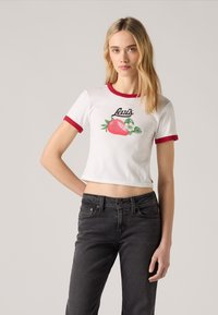 Valkoinen lyhythihainen t-paita punaisilla rengastakeilla, jossa on mansikan grafiikka ja musta "Levi's" -teksti, yhdistettynä tummiin farkkuihin.