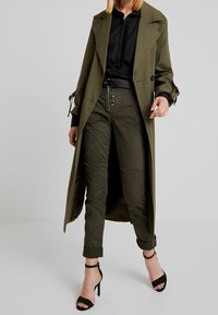 Lungo cappotto verde oliva con vita cinturata, abbinato a pantaloni olive con dettagli a bottoni e tacchi neri con strap. Textura liscia in tutto.