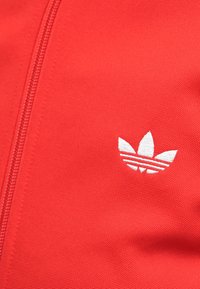 Sarkans audums ar rāvējslēdzēju pa kreisi un mazu balto izšūto Adidas trīsstūru logotipu labajā pusē.