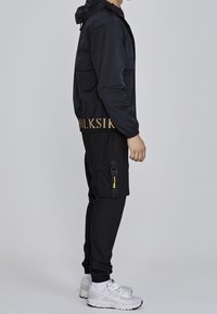 Black bomber jacket with hoodie, gold lettering at the hem, paired with black cargo pants featuring pockets and a yellow zipper pull, worn with white sneakers.
