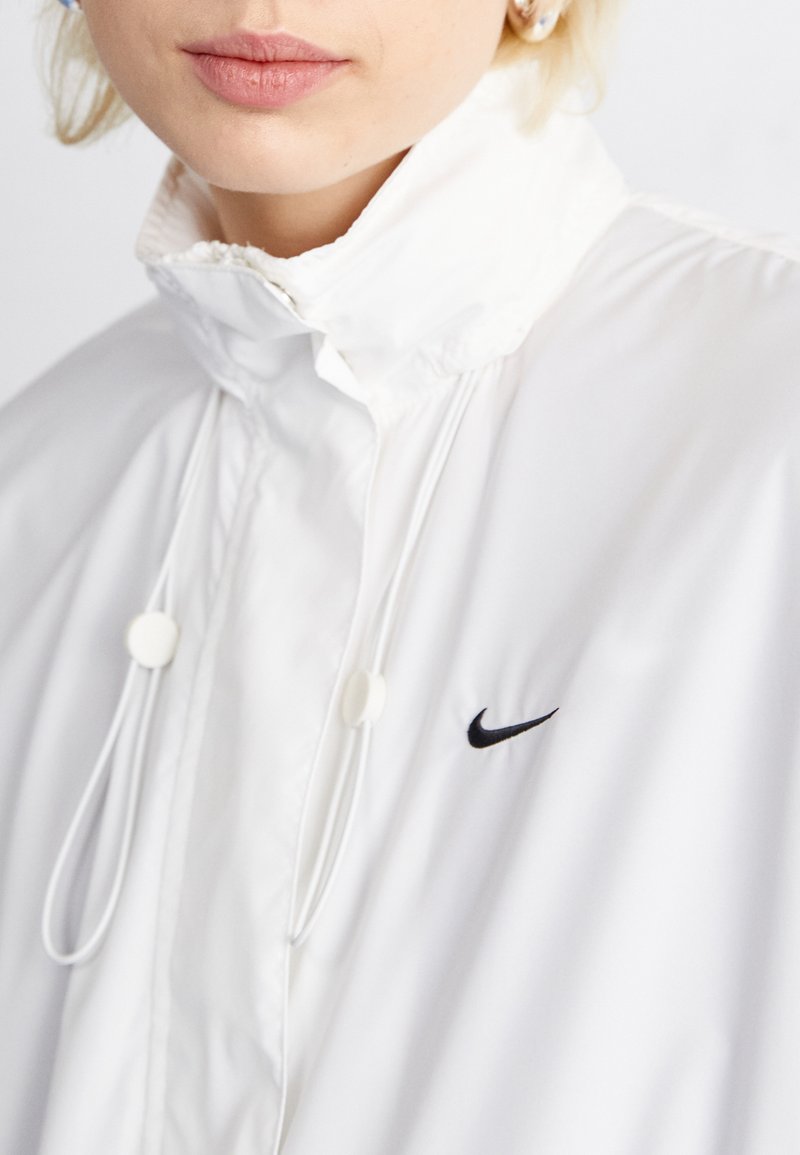 Witte windbreaker met hoge kraag, voorzien van het zwarte Nike-logo, verstelbare trekkoorden en sluitingen. Glad, lichtgewicht materiaal.